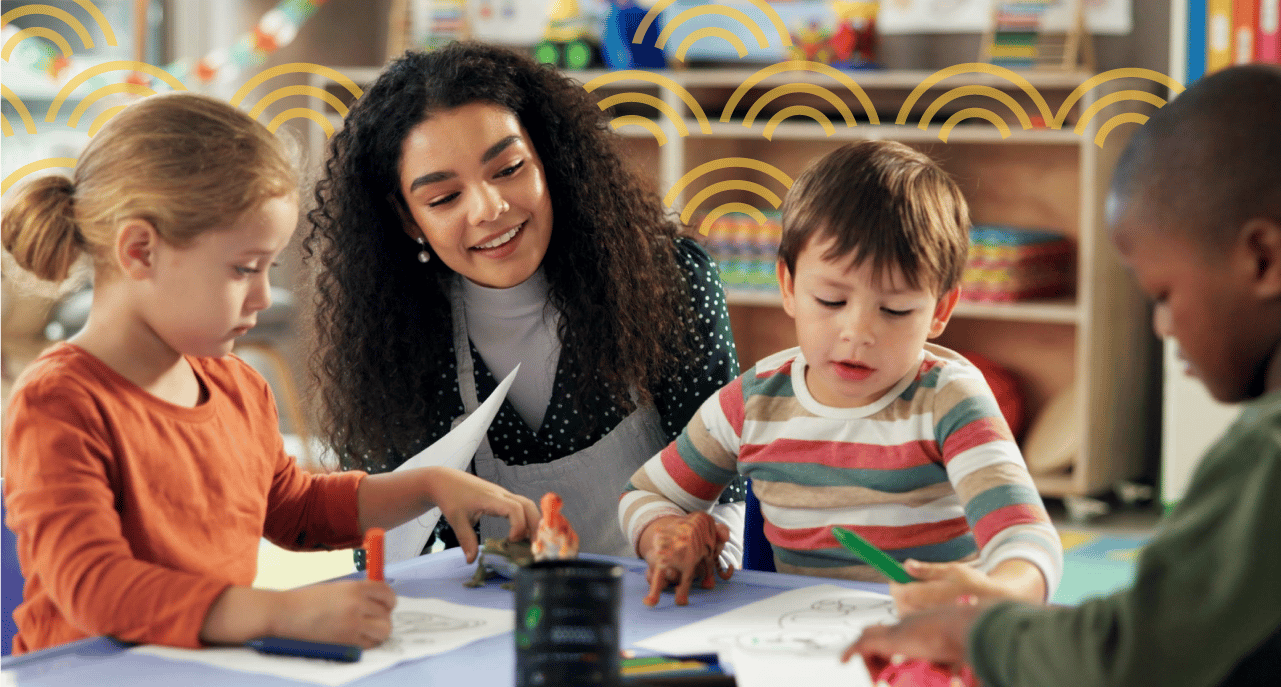 Three preschool-aged children sit at a table coloring and playing with toy animals while a female preschool teacher watches them