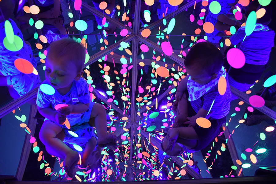 Babies sitting up interacting with brightly colored plastic circles hanging from the ceiling