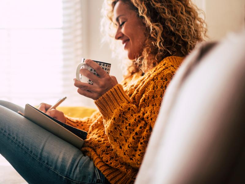 Woman in cozy sweater journaling on a sofa with a warm beverage.